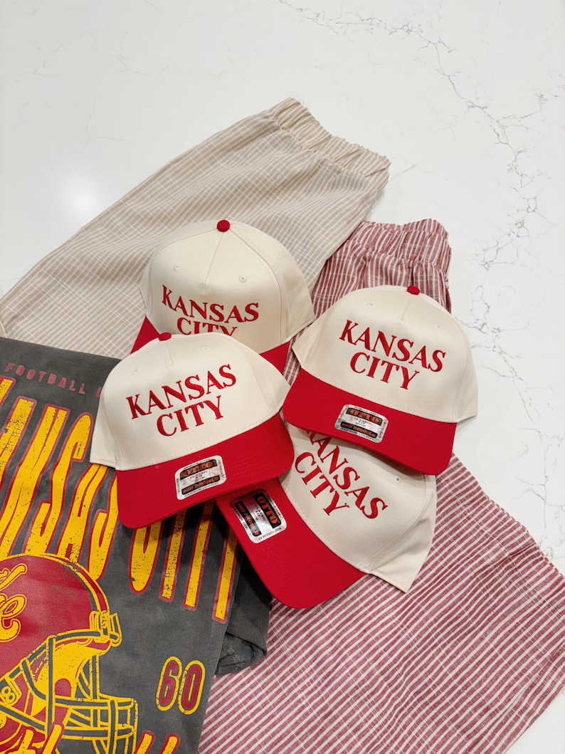 May include: Three white baseball caps with red brims and the text "Kansas City" in red lettering. The caps are on a striped fabric background.