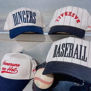 May include: Four baseball caps with white and navy blue color schemes. Two caps read "DINGERS" and "BASEBALL" in navy blue and black, respectively. Another cap says "Homeruns are Hot" in red. A baseball and glove are also present.