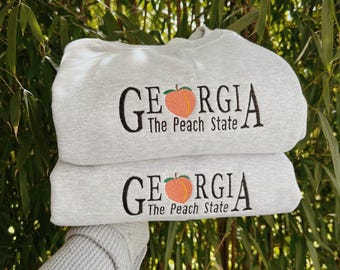 Könnte beinhalten: Zwei hellgraue Sweatshirts übereinander gestapelt. Jedes Sweatshirt hat den schwarzen Schriftzug "GEORGIA", eine Pfirsichgrafik und den Text "The Peach State" darunter.