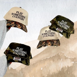 May include: Four baseball caps with a camouflage pattern and the text "PLAY SOMETHING COUNTRY" in white. The caps are in various positions, some with the camouflage pattern on the brim and others with the white fabric on the brim.