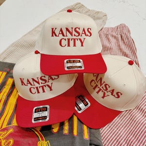 May include: Three white baseball caps with red brims. The caps have the text "KANSAS CITY" embroidered in red on the front. The caps are stacked on top of each other.