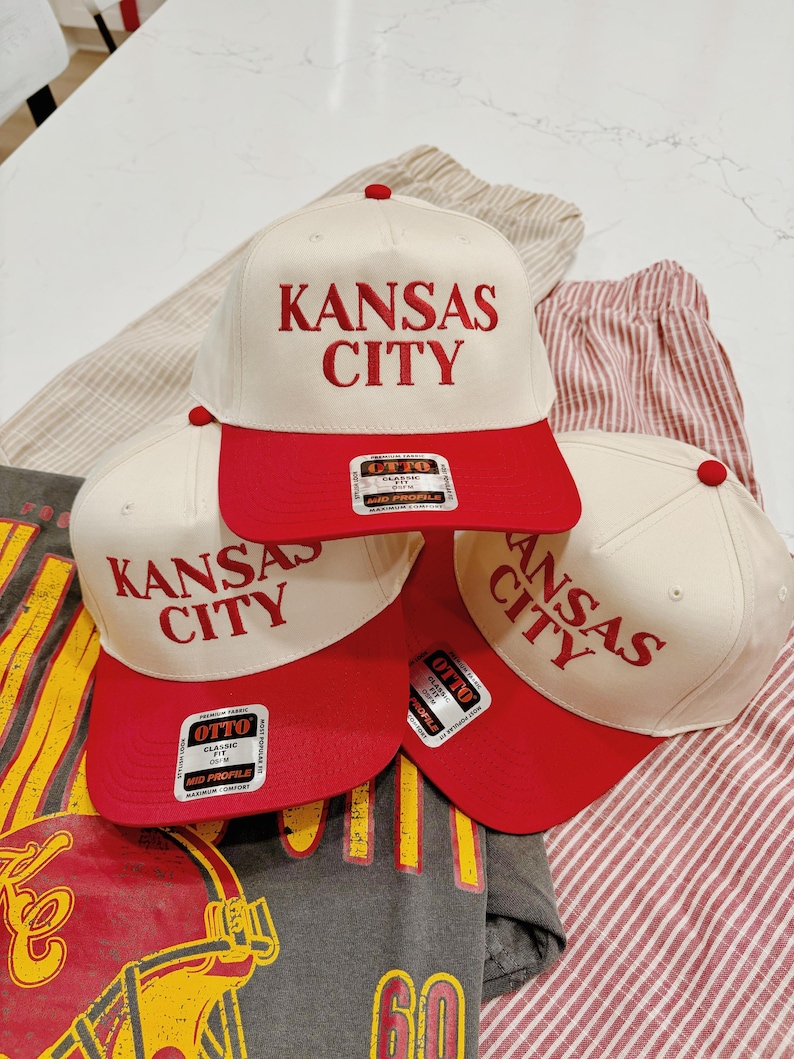 May include: Three white baseball caps with red brims. The caps have the text "KANSAS CITY" embroidered in red on the front. The caps are stacked on top of each other.