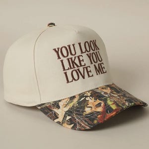 May include: A beige baseball cap with a camouflage brim. The front of the cap has the text "You look like you love me" embroidered in brown.