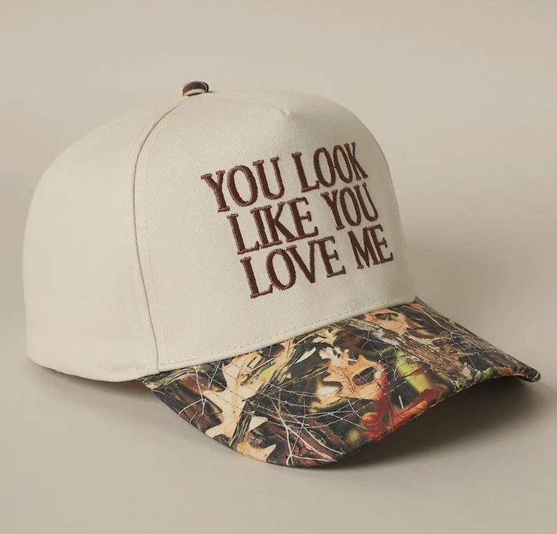 May include: A beige baseball cap with a camouflage brim. The front of the cap has the text "You look like you love me" embroidered in brown.
