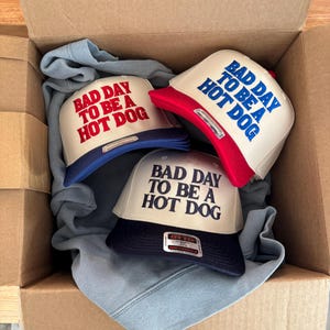 May include: Three baseball caps with the text "BAD DAY TO BE A HOT DOG" in red and blue lettering. The caps have a white front panel and a colored brim. They are inside a cardboard box with a blue garment.
