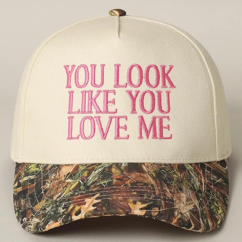 May include: A white baseball cap with a camouflage brim. The front of the cap has pink embroidered text that reads "You look like you love me."