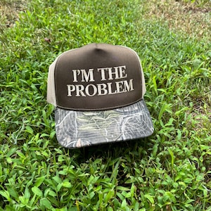 以下が含まれることがあります： 「I'M THE PROBLEM」の白い文字が入った茶色のトラッカーハット。迷彩柄のつばとベージュのメッシュバックが付いています。緑の草の上に置かれています。