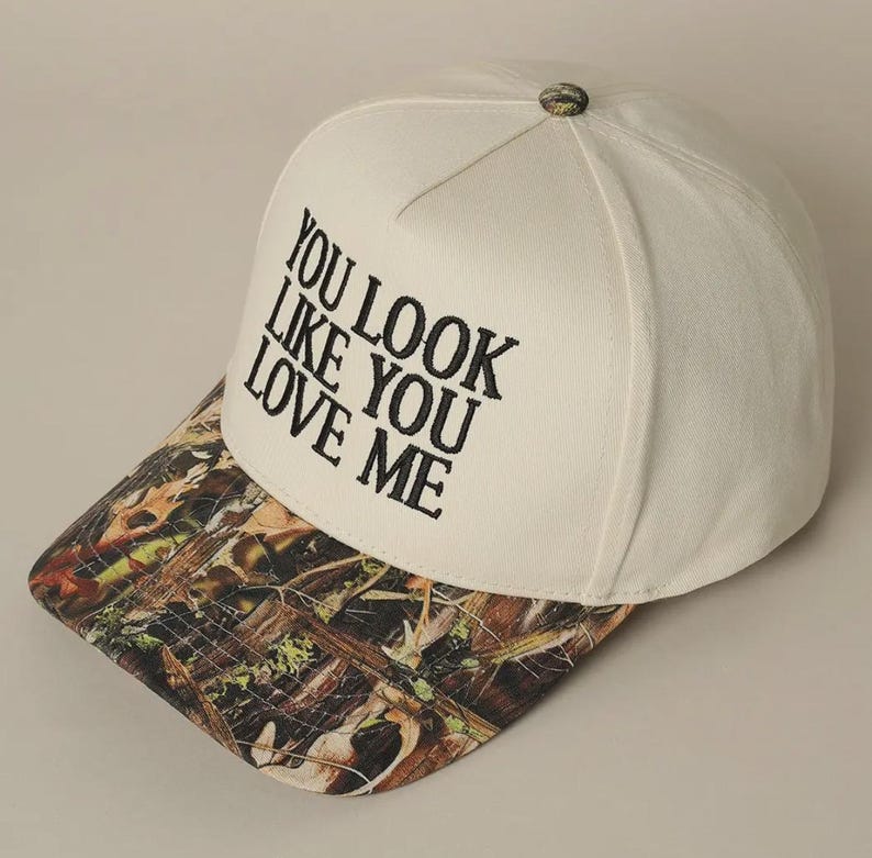May include: White baseball cap with a camouflage brim. The front of the cap has black embroidered text that reads "You look like you love me."
