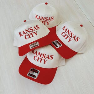 May include: Four white baseball caps with red brims. Each cap has the text "Kansas City" embroidered in red on the front.