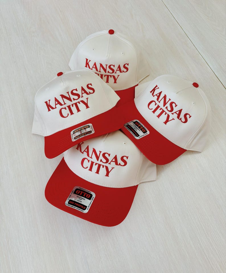 May include: Four white baseball caps with red brims. Each cap has the text "Kansas City" embroidered in red on the front.