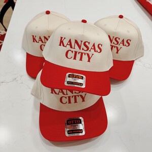 May include: Three white baseball caps with red brims. The caps have the text "KANSAS CITY" in red lettering. The caps have a label on the side that says "OTTO CLASSIC FIT OSFM MID PROFILE MAXIMUM COMFORT".