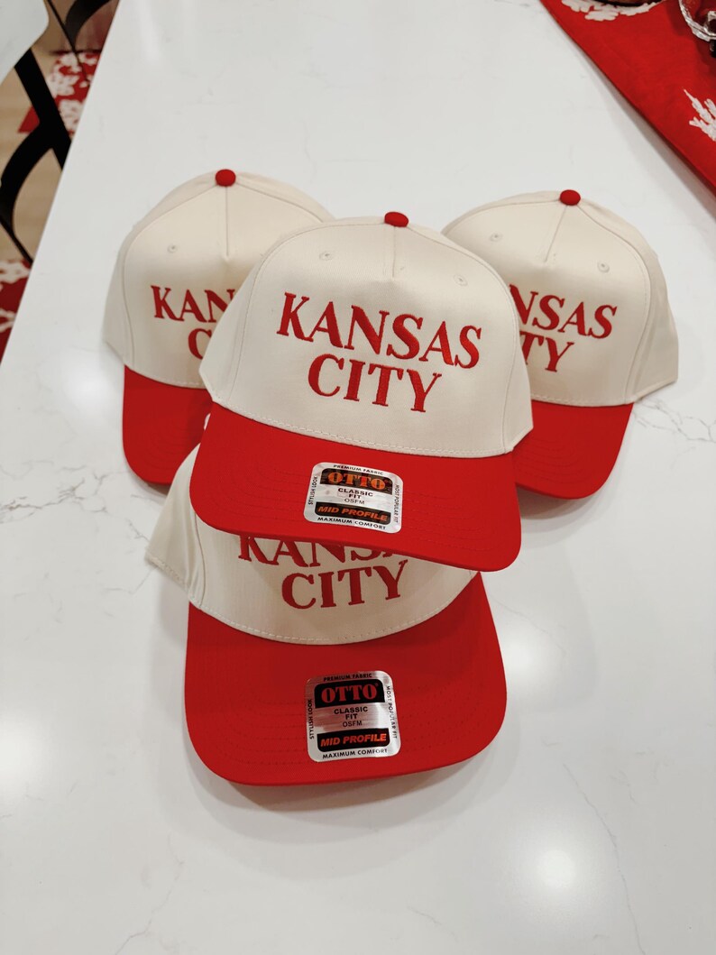 May include: Three white baseball caps with red brims. The caps have the text "KANSAS CITY" in red lettering. The caps have a label on the side that says "OTTO CLASSIC FIT OSFM MID PROFILE MAXIMUM COMFORT".