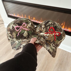 May include: Two camouflage baseball caps with embroidered bow designs. One cap features a pink bow, and the other has a red bow. The hats have a classic six-panel design and a curved brim.