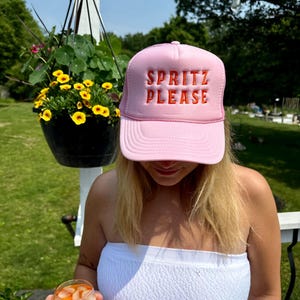 May include: A pink trucker hat with the words "SPRITZ PLEASE" embroidered in orange. The hat is worn by a person. The background includes a hanging flower basket and greenery.