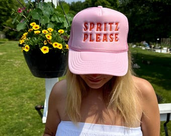 May include: A pink trucker hat with the words "SPRITZ PLEASE" embroidered in orange. The hat is worn by a person. The background includes a hanging flower basket and greenery.
