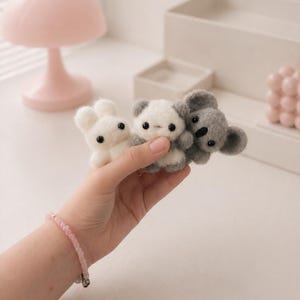 May include: Three small, handmade felt animal figurines: a white bunny, a panda, and a grey koala. The bunny and panda are white with black eyes, while the koala is grey. The figurines are held in a hand, with a pink beaded bracelet on the wrist.