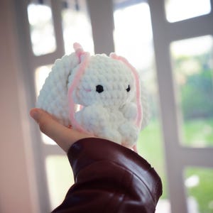 Op de afbeelding: Een handgemaakt wit gehaakt konijn met roze accenten. Het konijn heeft zwarte ogen en lange, slappe oren met roze garendetails. Het pluche speelgoed wordt omhoog gehouden tegen een vage achtergrond van een raam en groen.