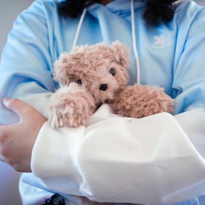 May include: A light brown teddy bear with black eyes and a black nose is held in a light blue hoodie with a white star design. The bear is nestled in a white fabric.