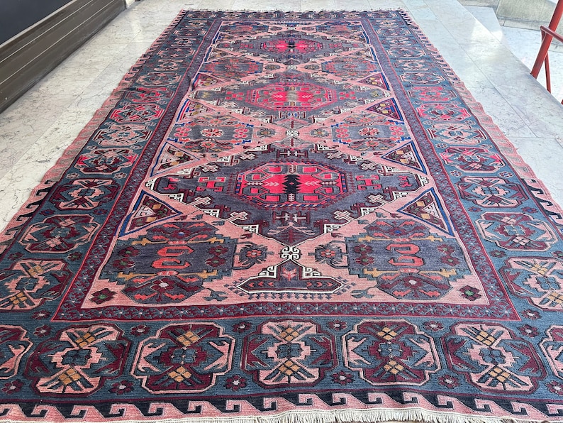 May include: A vintage Persian rug with a traditional geometric pattern in shades of pink, blue, red, and black. The rug has a fringed edge and is made of wool.