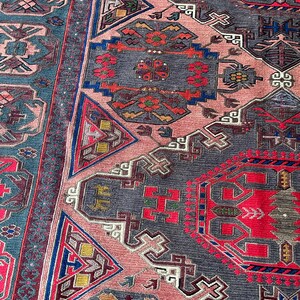 May include: A close-up of a traditional rug with intricate geometric patterns in shades of red, blue, pink, and gray. The rug features a repeating design of triangles, squares, and other shapes.