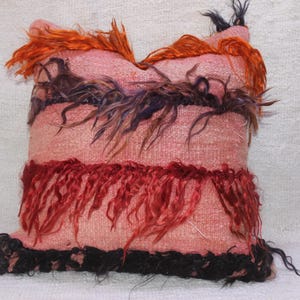 May include: A pink throw pillow with red, orange, and black fringe. The pillow has a woven texture and is decorated with colourful strands of yarn.