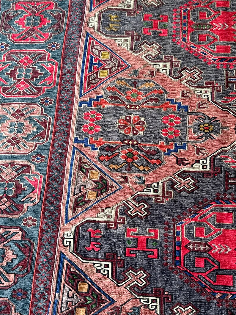 May include: A close-up of a traditional rug with intricate geometric patterns in shades of red, blue, pink, and gray. The rug features a repeating design of triangles, squares, and crosses.