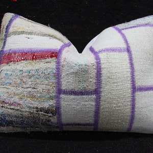May include: Rectangular decorative pillow with a patchwork design. The pillow features a white background with purple stitching and accents. The patchwork includes various colors like red, blue, and yellow, creating a textured appearance. The pillow is likely for home decor.