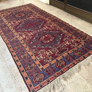 May include: A vintage rug with a geometric pattern in shades of red, blue, and beige. The rug has a fringe border and is woven with intricate details.