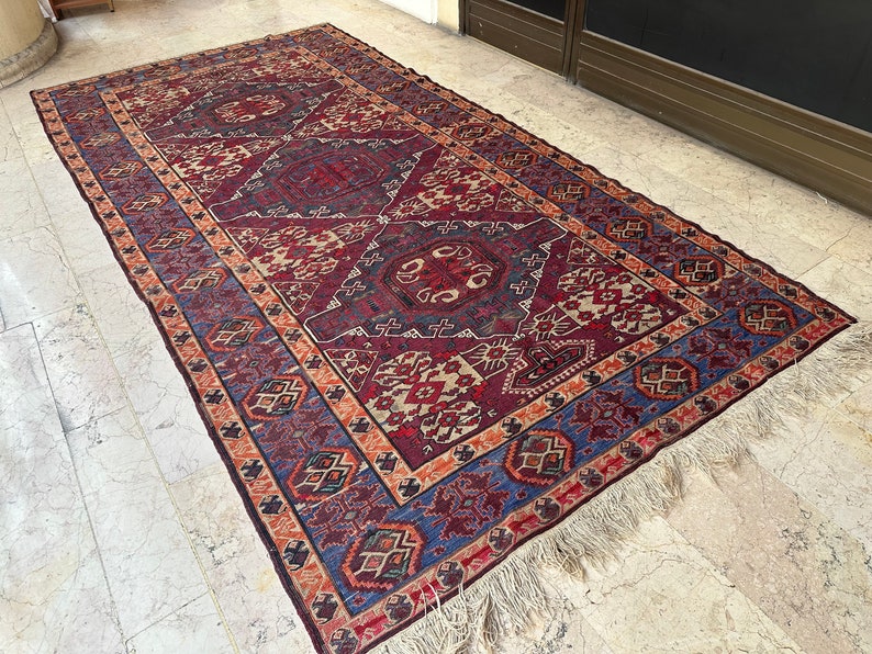 May include: A vintage rug with a geometric pattern in shades of red, blue, and beige. The rug has a fringe border and is woven with intricate details.