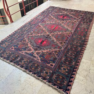 May include: A vintage rug with a pink, blue, and green geometric pattern. The rug has a fringe border and is laid out on a tiled floor.