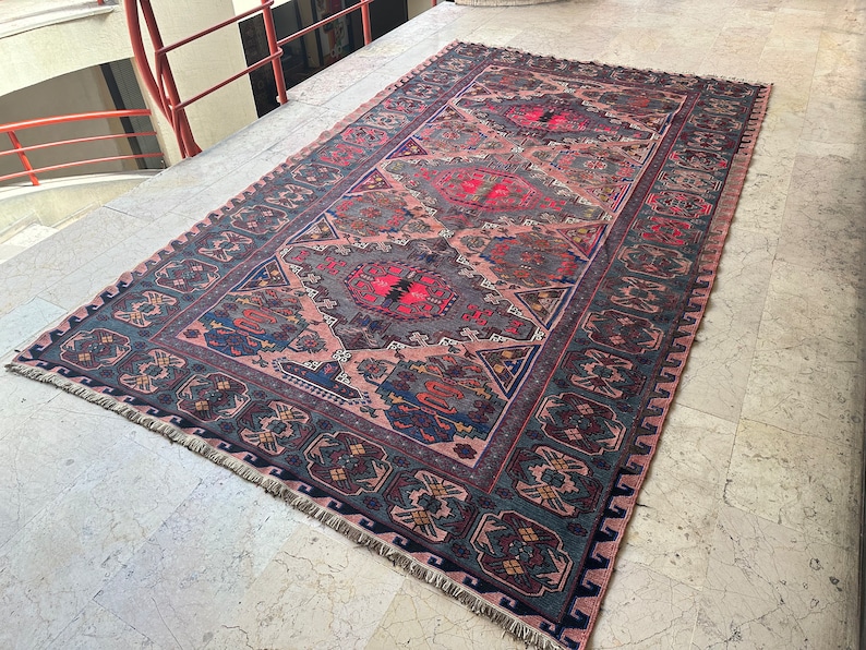 May include: A vintage rug with a pink, blue, and green geometric pattern. The rug has a fringe border and is laid out on a tiled floor.