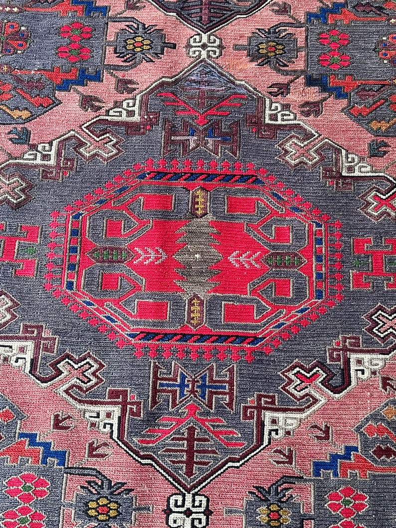 May include: A close-up of a pink and gray rug with intricate geometric patterns in shades of red, blue, and green. The rug has a traditional design with a central medallion and repeating motifs.
