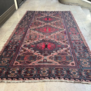 May include: A vintage rug with a pink background and intricate geometric patterns in shades of blue, red, and black. The rug has a fringed edge.