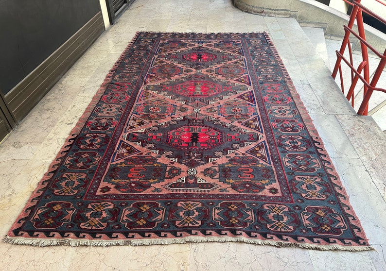 May include: A vintage rug with a pink background and intricate geometric patterns in shades of blue, red, and black. The rug has a fringed edge.