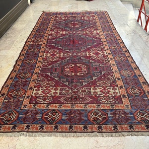 May include: A traditional Persian rug with a red, blue, and cream color scheme. The rug features a geometric pattern with intricate details. The rug has a fringe border.