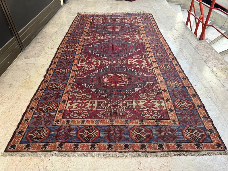 May include: A traditional Persian rug with a red, blue, and cream color scheme. The rug features a geometric pattern with intricate details. The rug has a fringe border.