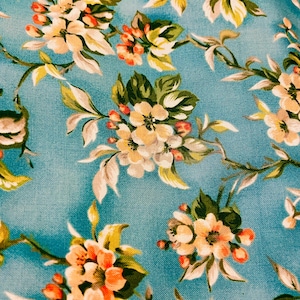 May include: Close-up of a floral fabric with a teal background. The pattern features clusters of cream-colored flowers with orange centers and green leaves. Delicate vines and branches weave throughout the design.