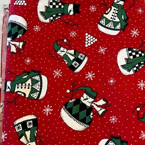 May include: Red fabric with a white snowflake pattern and a repeating pattern of snowmen wearing green and white hats, scarves, and sweaters. The fabric has the text "www.northcott.net" and "100% Cotton" printed on it.