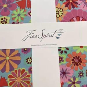 May include: A stack of fabric squares with a floral pattern in shades of red, pink, orange, yellow, and green on a light blue background. The fabric is wrapped with a white band that reads "Free Spirit" in cursive.