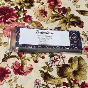 May include: A package of "Bricolage" 10 Karat Crystals by Kaye England rests on a floral patterned fabric. The packaging has a navy blue and white design with the brand name and product details. The fabric features large, detailed flowers in shades of red, pink, and cream.