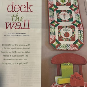 May include: A festive wall hanging or table runner with a red, green and white patchwork design. The runner features three embroidered ornaments with a butterfly and a bell design. The text "deck the wall" is printed on the page.