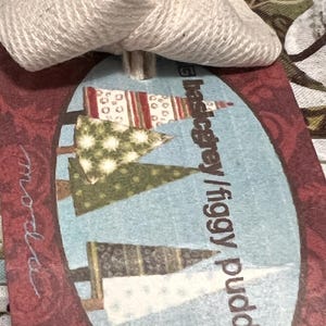 May include: A red and white fabric tag with a blue oval in the centre. The oval features a design of three stylised Christmas trees with a white background. The text "badgergrey / figgy pudding" is printed in brown on the tag.