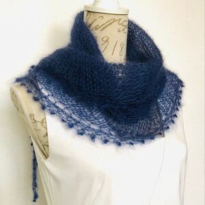 May include: A dark blue, hand-knitted mohair scarf with a loose, open-weave texture. The scarf features a delicate, scalloped edge with small, fuzzy bobble details along the border, draped around the neck of a white mannequin torso.