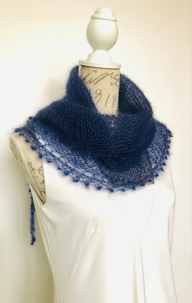 May include: A dark blue, hand-knitted mohair scarf with a loose, open-weave texture. The scarf features a delicate, scalloped edge with small, fuzzy bobble details along the border, draped around the neck of a white mannequin torso.