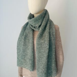 Peut inclure: Une longue écharpe tricotée de couleur vert sauge doux avec un aspect texturé et duveteux. L'écharpe est drapée autour du cou d'un mannequin, qui porte également un pull à manches longues beige clair et une jupe blanche unie.