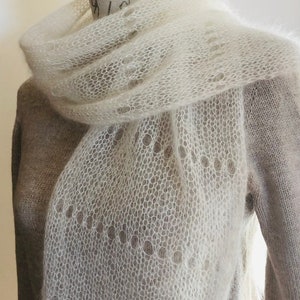 May include: A long, off-white, semi-sheer knitted scarf featuring a delicate, open-work lace pattern with circular eyelet details. The scarf is crafted from a soft, fuzzy yarn and is draped around the neck of a mannequin wearing a beige sweater.