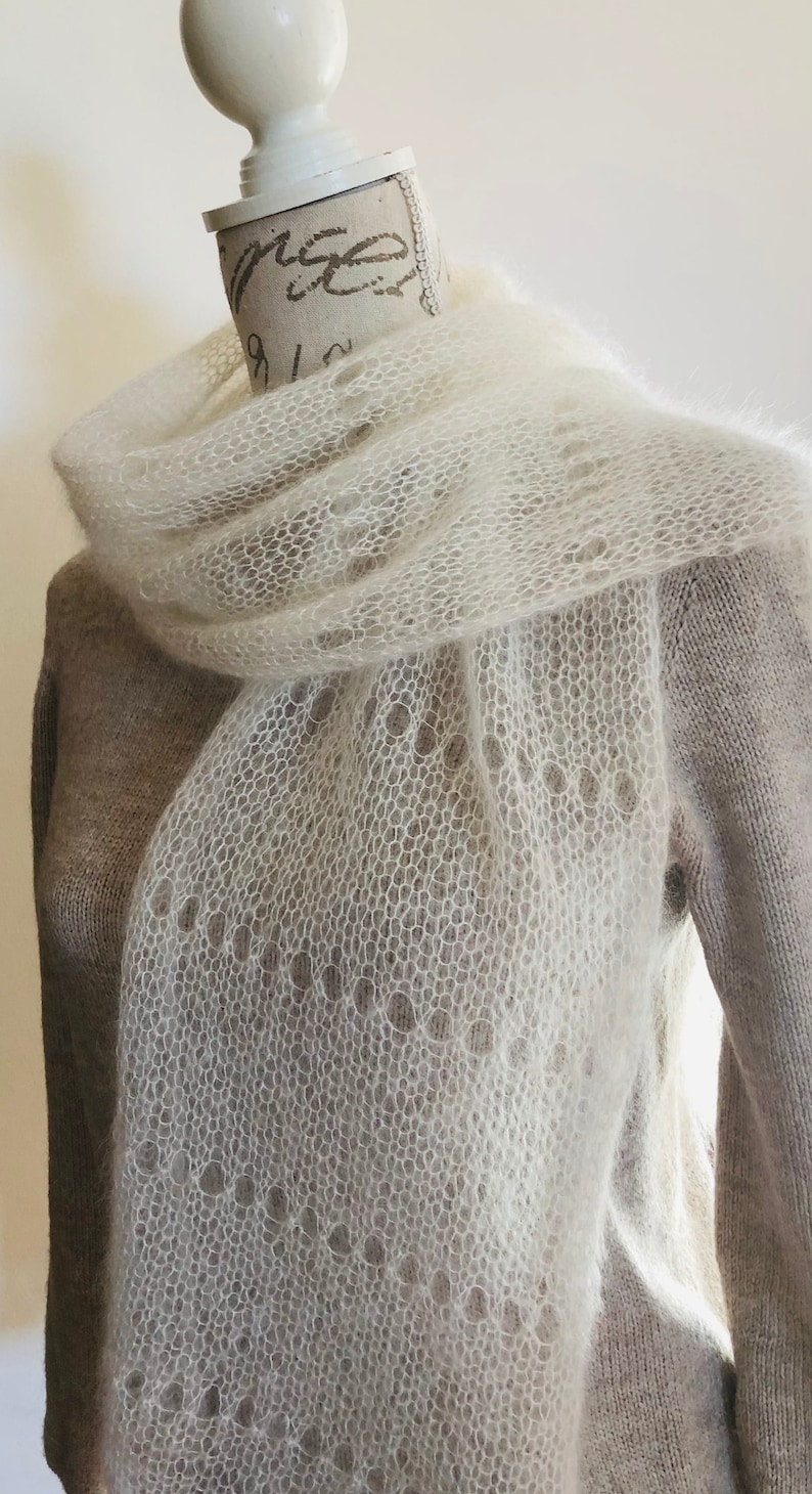 May include: A long, off-white, semi-sheer knitted scarf featuring a delicate, open-work lace pattern with circular eyelet details. The scarf is crafted from a soft, fuzzy yarn and is draped around the neck of a mannequin wearing a beige sweater.