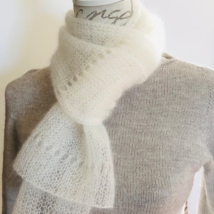 May include: A white, airy, open-knit scarf with a delicate, lacy texture draped around the neck of a mannequin wearing a light beige, long-sleeved, fine-knit sweater. The scarf features a subtle, repeating pattern of small, circular eyelet holes.