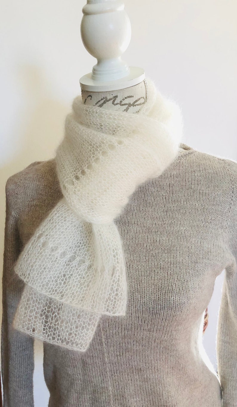 May include: A white, airy, open-knit scarf with a delicate, lacy texture draped around the neck of a mannequin wearing a light beige, long-sleeved, fine-knit sweater. The scarf features a subtle, repeating pattern of small, circular eyelet holes.
