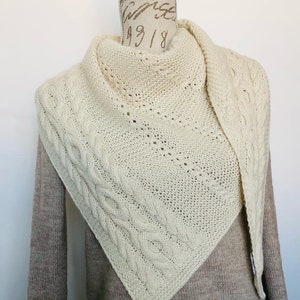 Puede incluir: Chal de punto color crema con diseño de punto trenzado y forma triangular. El chal está drapeado sobre un maniquí, mostrando su textura y patrón intrincado. El chal está hecho de un material suave y cálido.